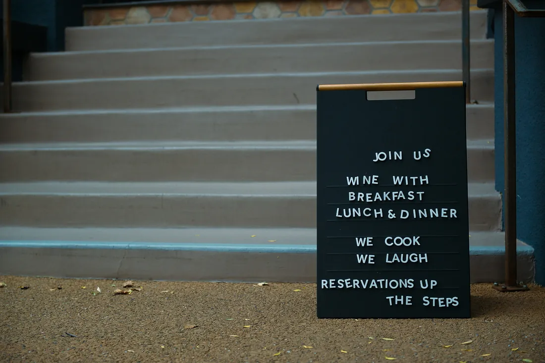 A letterboard sign at the foot of a flight of steps reading: join us, wine with breakfast lunch and dinner, we cook we laugh, reservations up the steps.