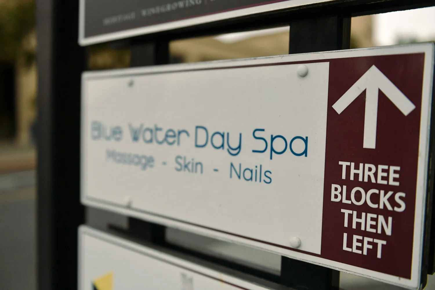 A directional sign for a day spa listing massage, skin and nails, beside an arrow marked three blocks then left.