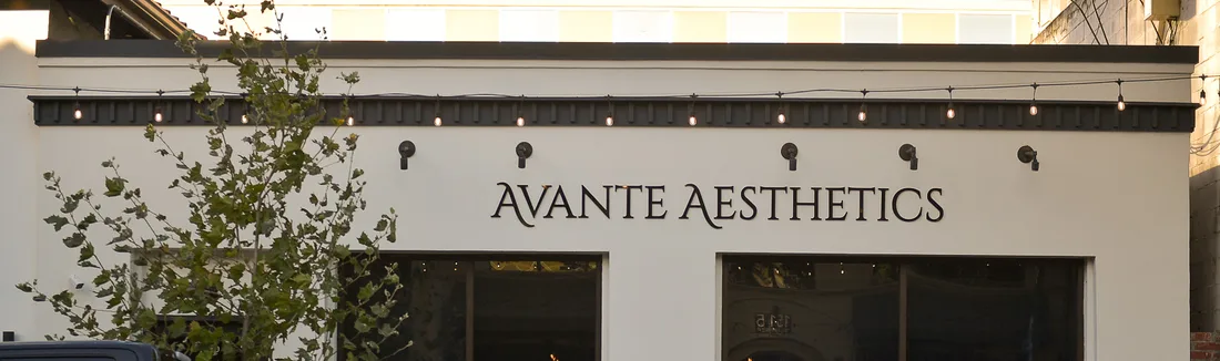 The upper facade of a Walnut Creek aesthetics clinic: lettering reading Avante Aesthetics below a run of festoon lights and wall sconces, with a young tree in front.