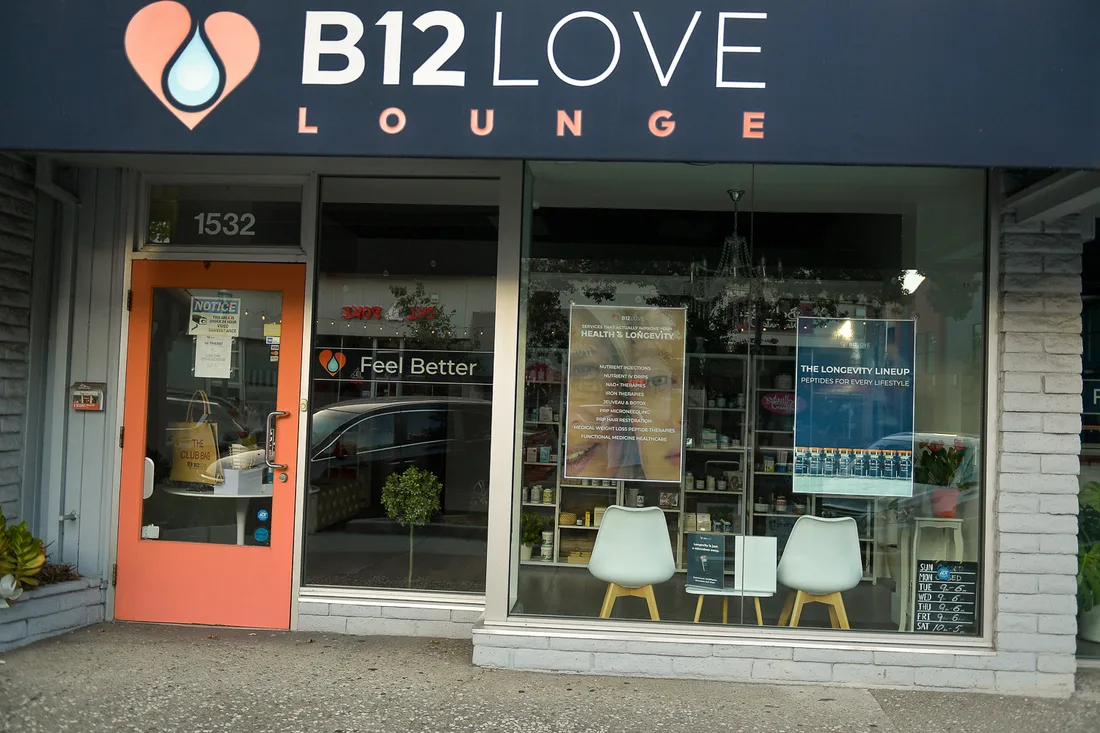 The B12 Love Lounge storefront at 1532: a navy sign, an orange door, and windows listing health and longevity services, with a small chalkboard of opening hours at the right.