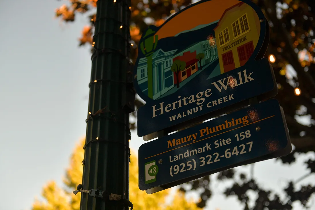 A Heritage Walk marker in Walnut Creek at dusk, illustrated with a fire station and storefronts, above a plaque reading Mauzy Plumbing, Landmark Site 158, and a phone number.