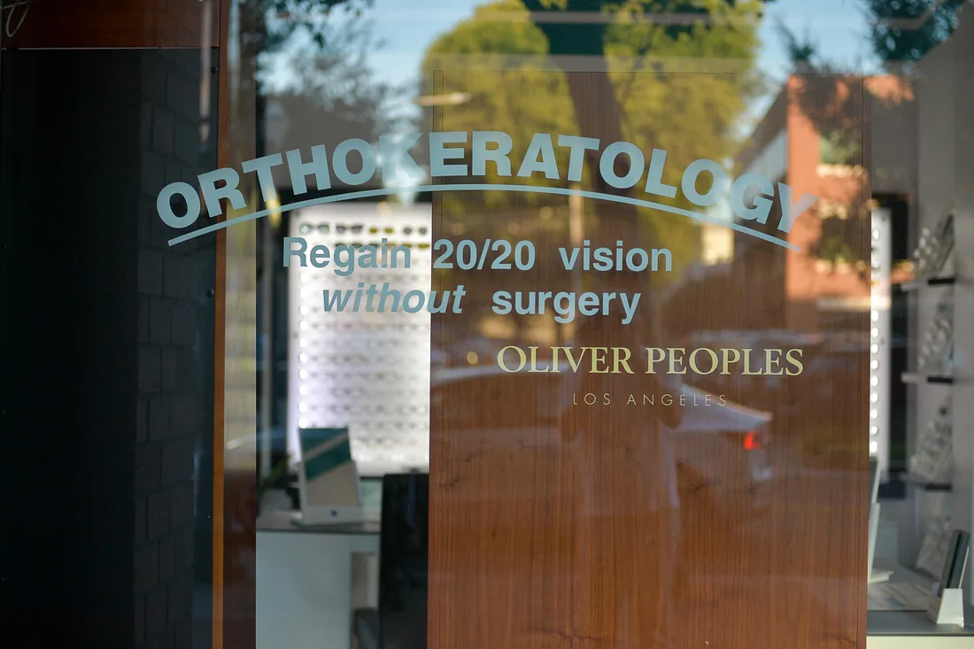 An optician's window lettered Orthokeratology, regain 20/20 vision without surgery, with trees and a brick building reflected in the glass and spectacle displays visible inside.