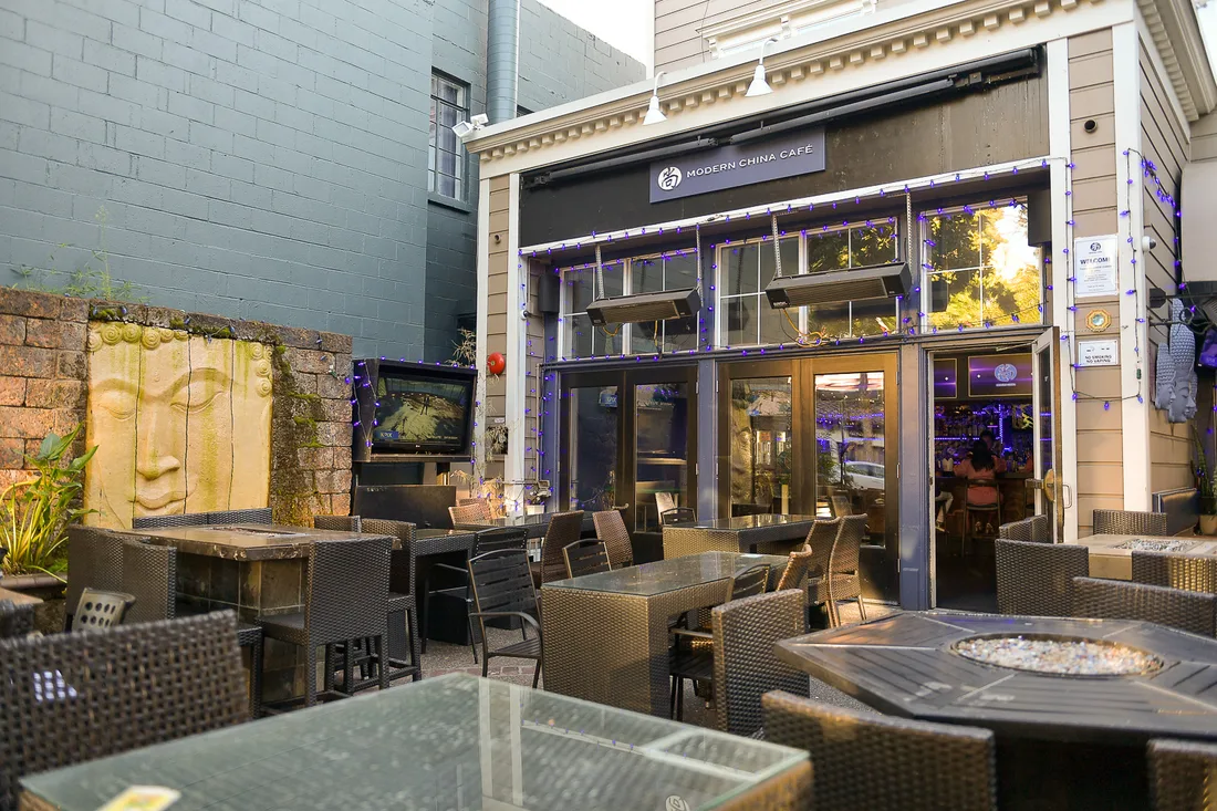 An empty restaurant courtyard in Walnut Creek at dusk: wicker chairs and glass-topped tables, string lights along the eaves, and a carved stone face set into the wall.