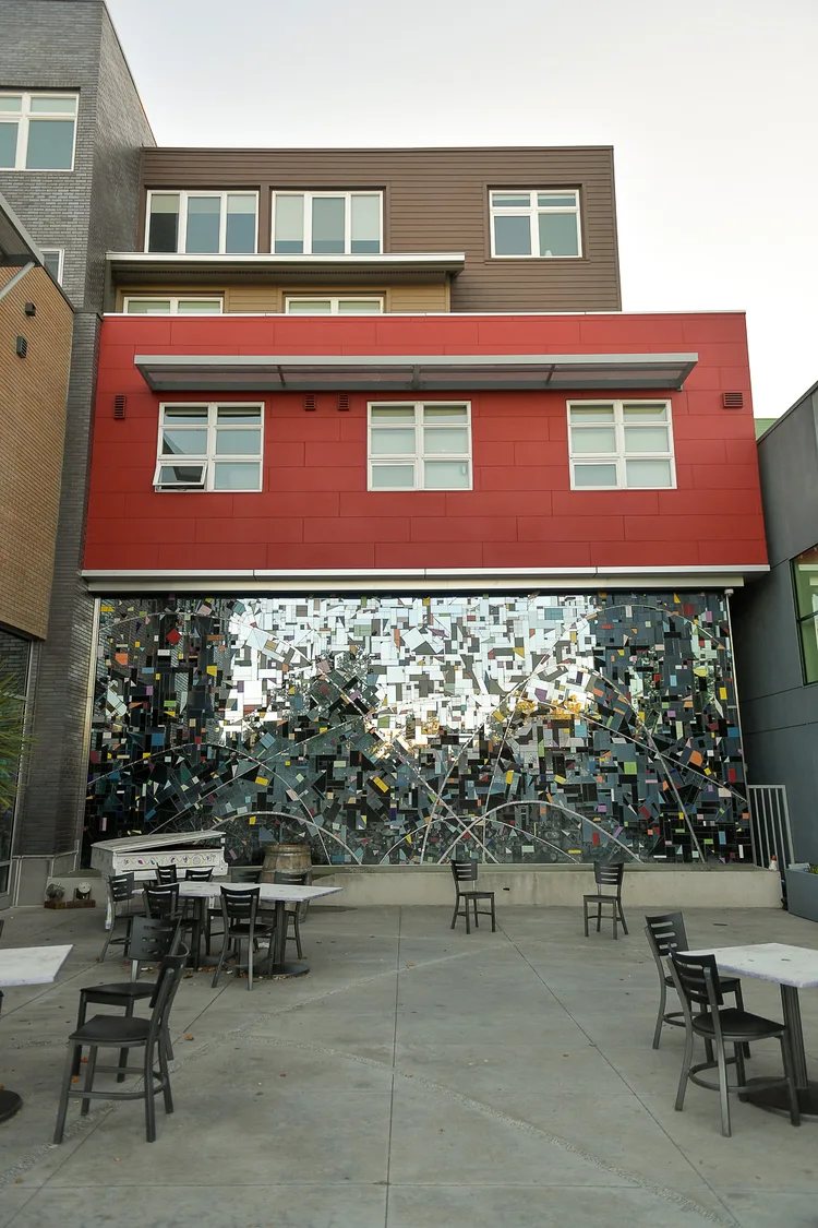 A public plaza in Walnut Creek: bistro tables and chairs on concrete in front of a large wall mosaic of mirrored and coloured tiles, under a red-panelled building.